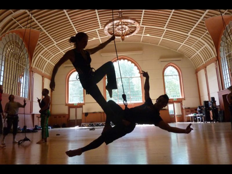 An EADF participant is suspended at the hips low to the ground in an elegant shape, while their partner stands on their hip holding the rope