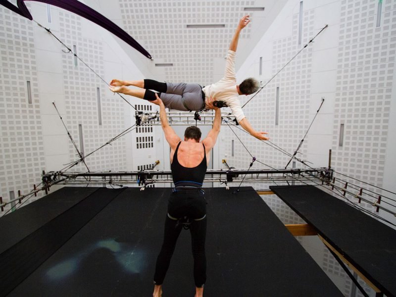 An aerialist appears to stand on the floor, effortlessly lifting another aerialist over their head, the ease in their bodies defies gravity
