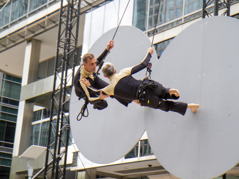 Vertical dance duet, Why?, performed on a grey wall made of circles, rigged in front of an urban building, at GDIF 2021