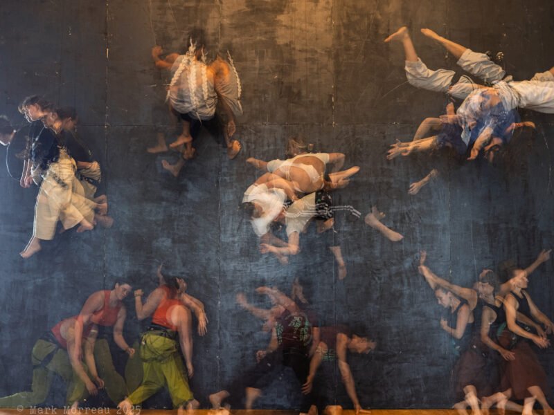 Against a black abseil wall a group of vertical dancers wearing brightly coloured clothing move together, some on the floor, some in the air making different shapes. The shot shows multiple exposures and is reminiscent of an oil painting