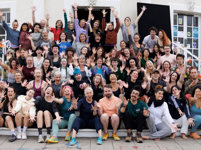 A group of people gather on the steps in front of a semi circlular building facade. Some sit and some stand but all are laughing, shouting and gesticulating towards the camera